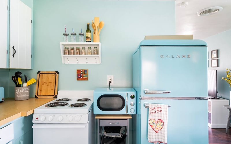 Retro-style kitchen with turquoise walls, white stove, blue microwave, Galanz refrigerator, and open shelving with spice jars and utensils.