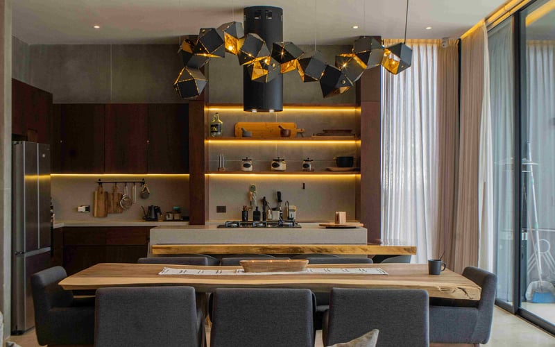 Modern kitchen and dining area with dark cabinetry, gold accent lighting, geometric pendant lights, wood dining table, and gray upholstered chairs