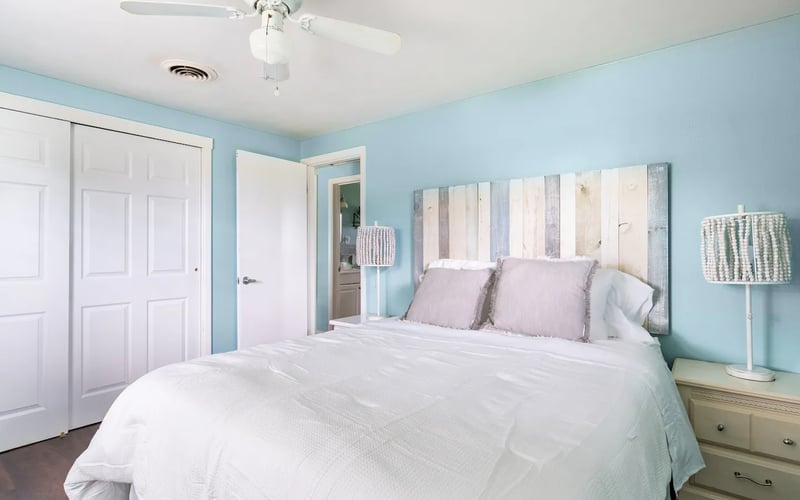 Bedroom with light blue walls, white ceiling fan, queen bed with white bedding and purple pillows, wooden nightstand, and white closet doors.